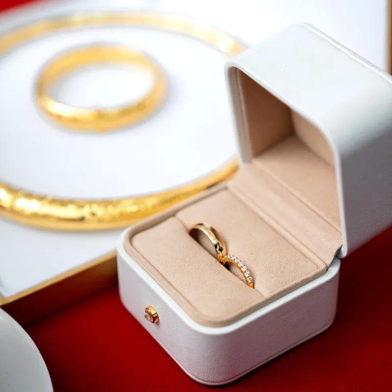 Sepasang cincin emas di dalam kotak perhiasan putih, dengan gelang emas di latar belakang